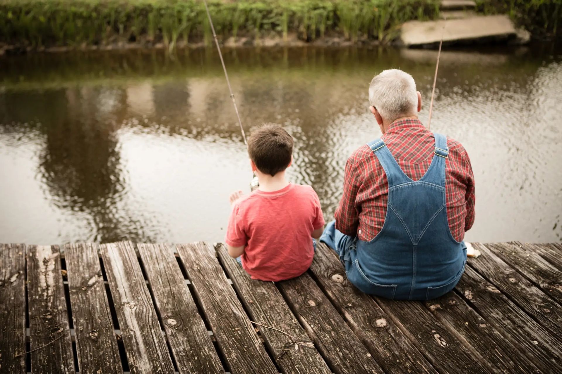 Two people sit on a weathered dock, fishing beside a calm pond.
