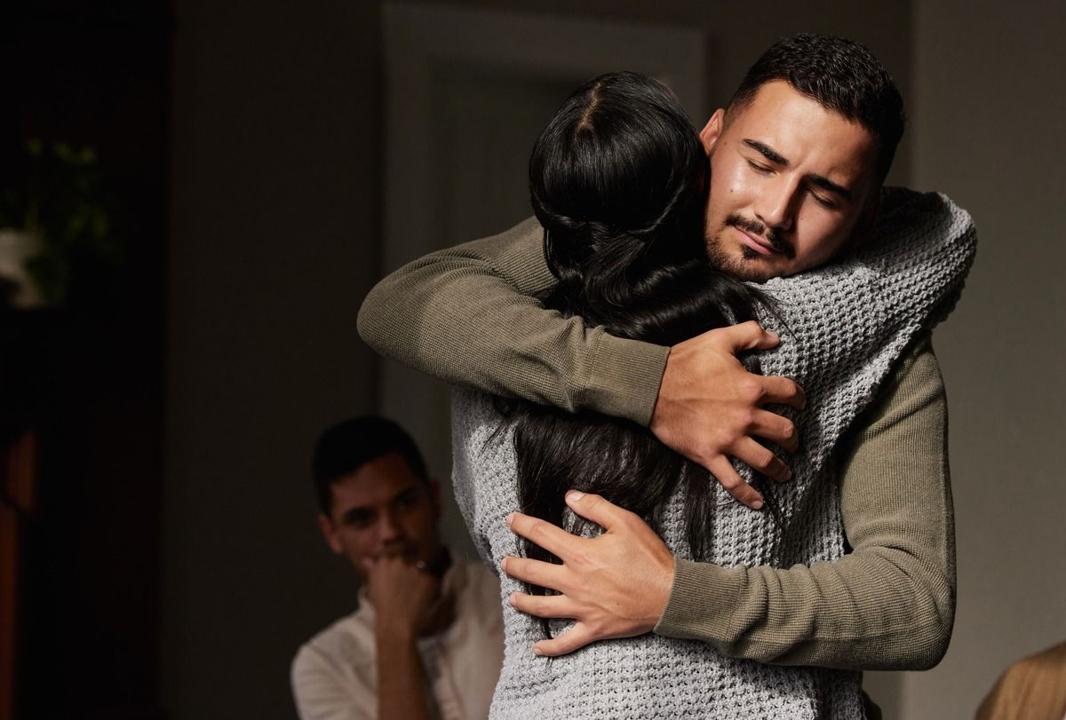 Two people hugging tightly indoors, with a man watching in the background.