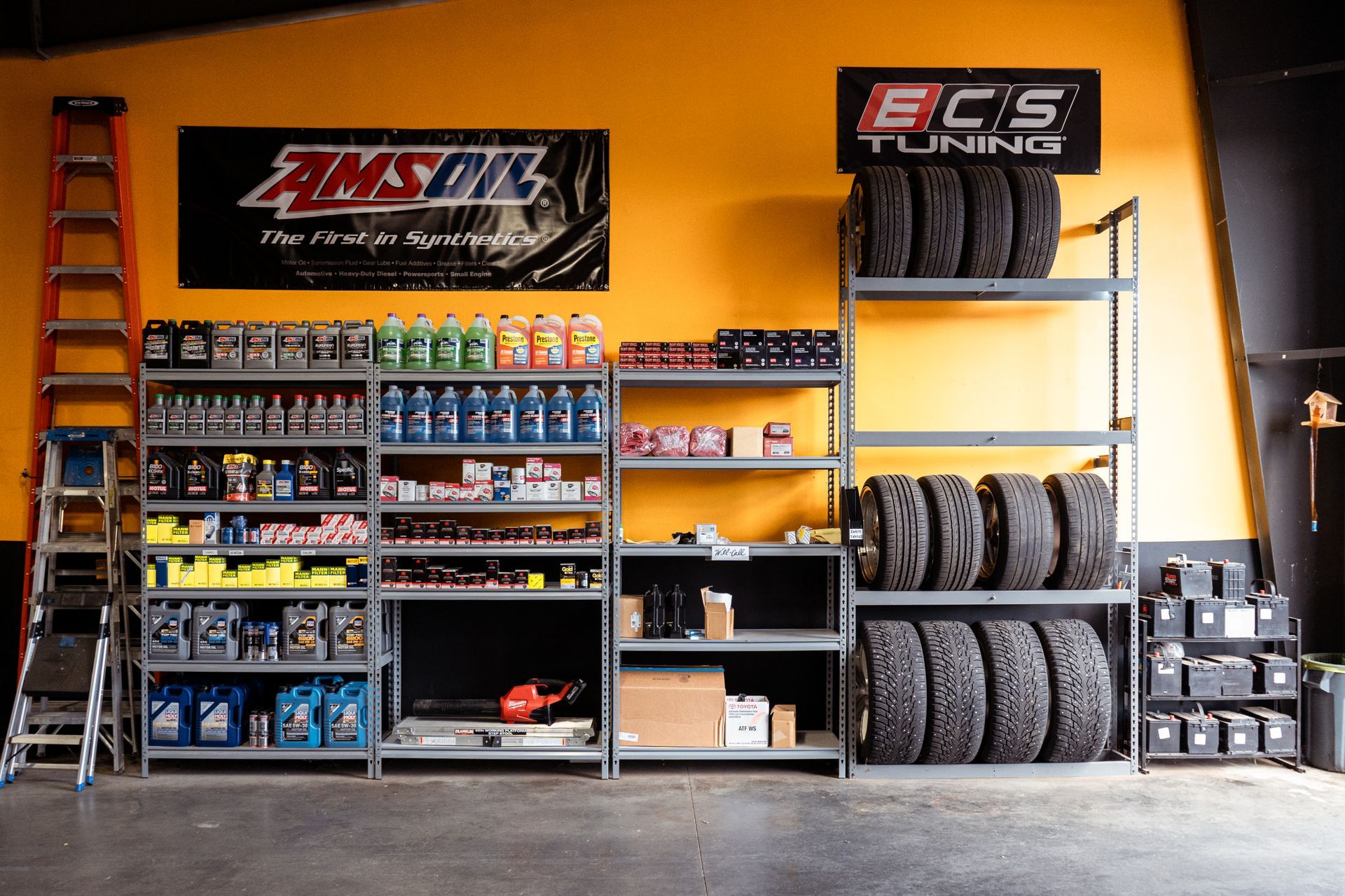 Auto shop wall with ECS Tuning signs, shelves of parts and fluids, and stacks of tires on a yellow background | VP Automotive LLC