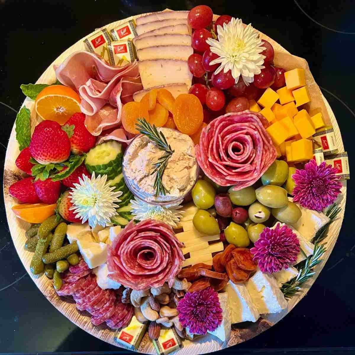 A plate of food with meat , cheese , fruit and flowers on a table.
