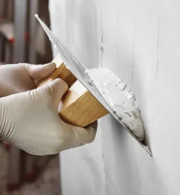 Des mains gantées de blanc tenant une truelle, appliquant de l'enduit sur un mur blanc.