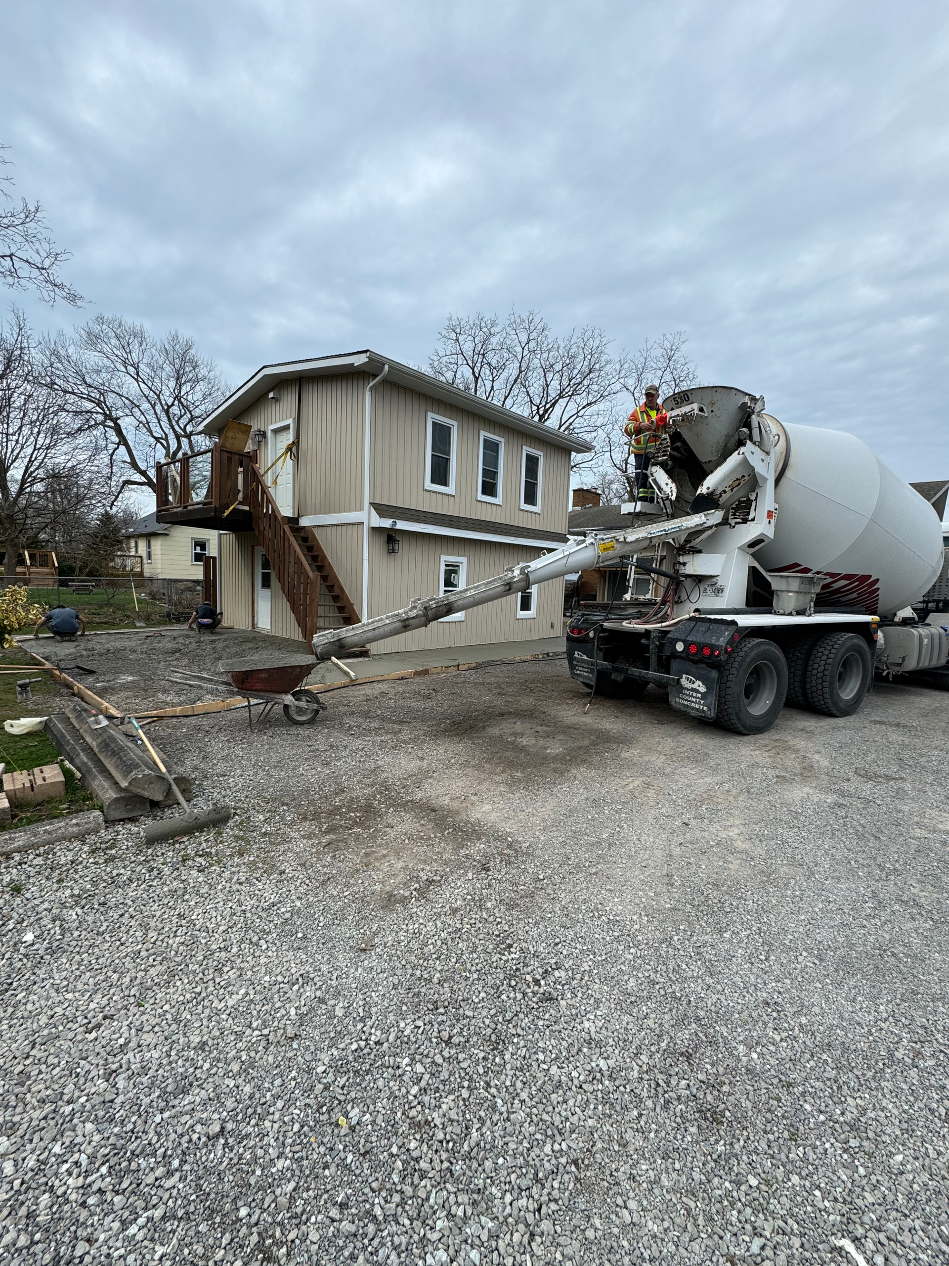 A concrete mixer truck is pouring concrete into a gravel driveway in front of a house.