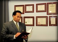 A person in a suit and tie reads a document in front of a wall decorated with multiple framed professional certificates.