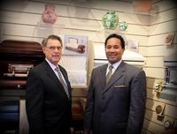 Two men in suits stand smiling in front of a wall display featuring vases and burial caskets.