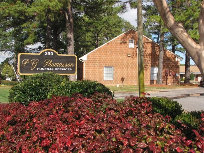 A sign for P.G. Thomasson Funeral Services stands in front of a brick building surrounded by trees and bushes.