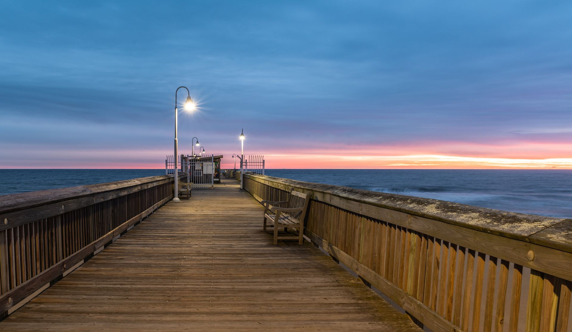 A wooden pier stretches toward the ocean at dusk, with glowing lights and a sunset sky over the horizon.