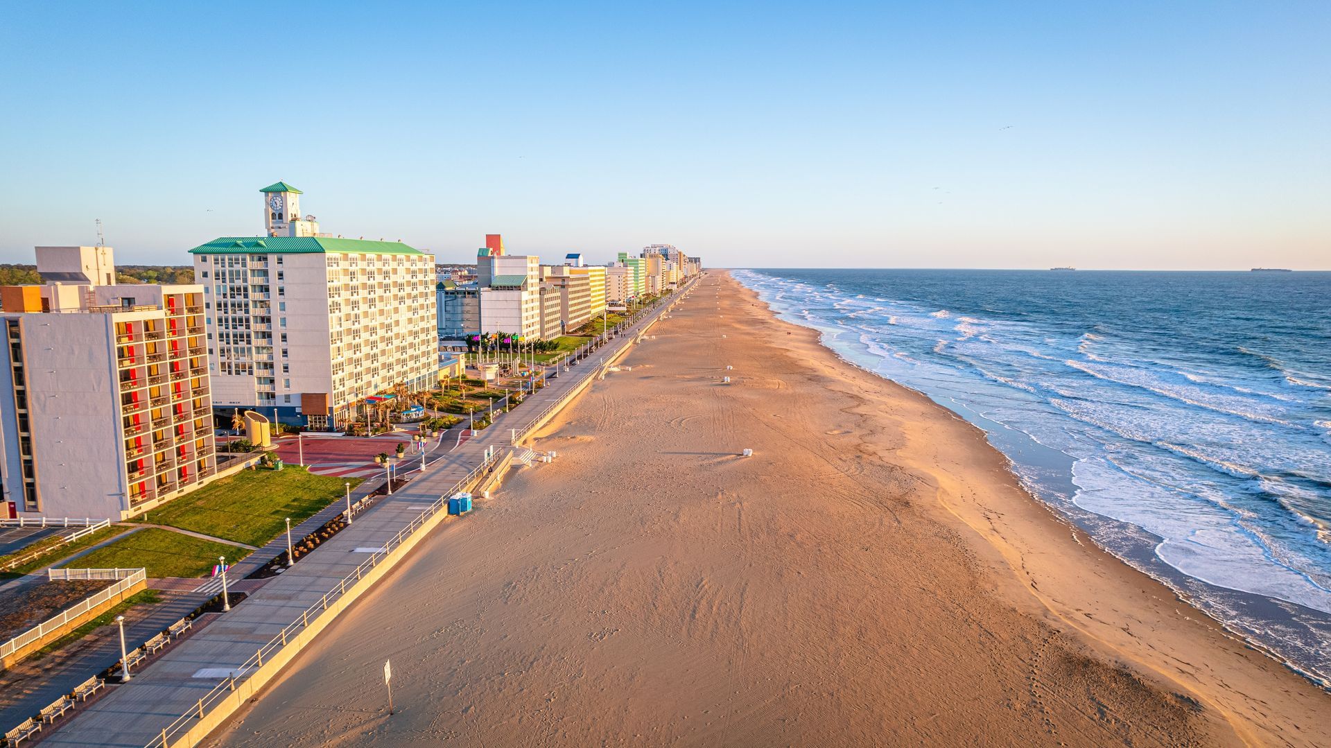 An aerial view of a sandy beach alongside a boardwalk, tall hotels, and the blue ocean at sunset.