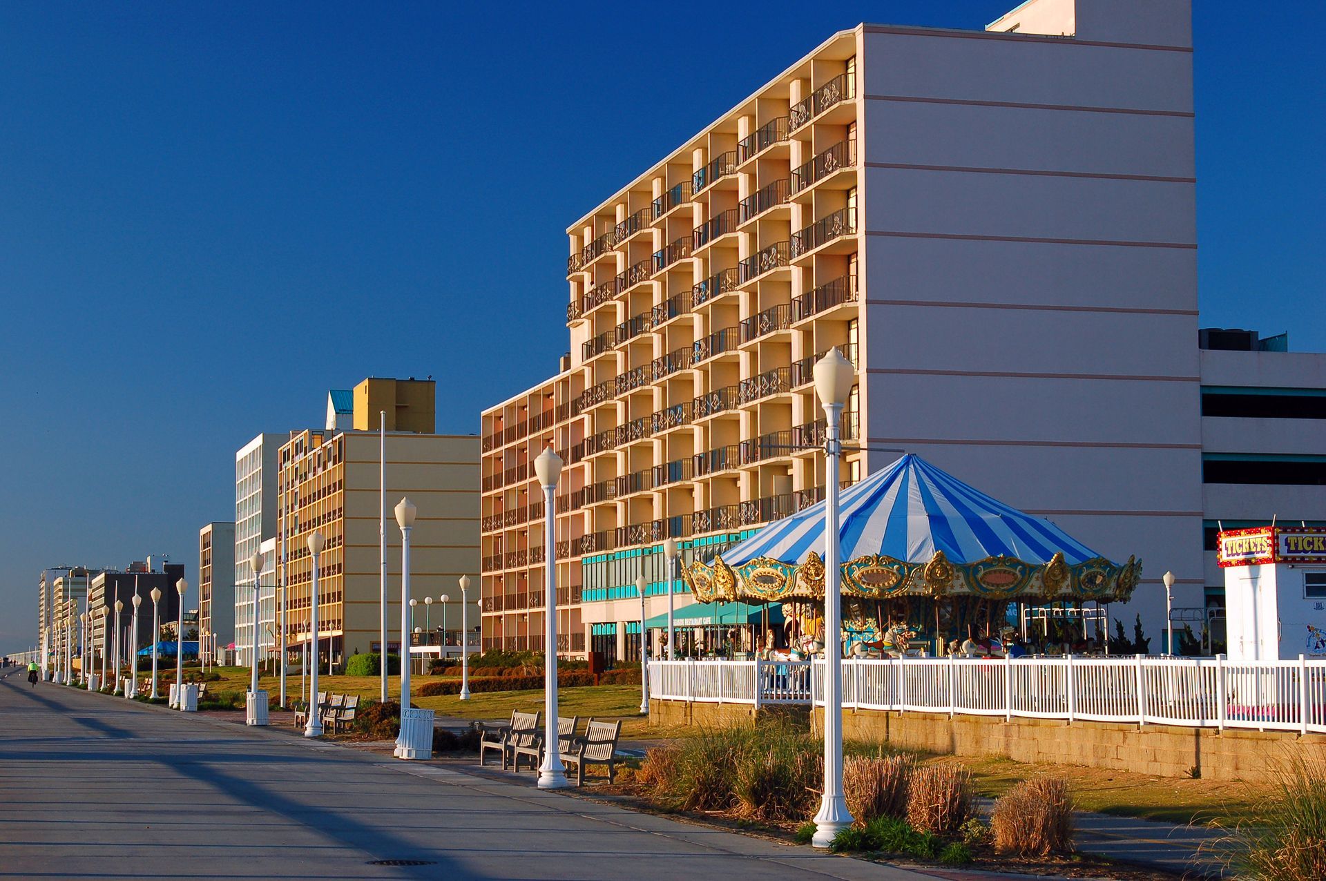 A wide boardwalk stretches along a row of beachfront hotels under a clear blue sky, featuring a carousel in the foreground.