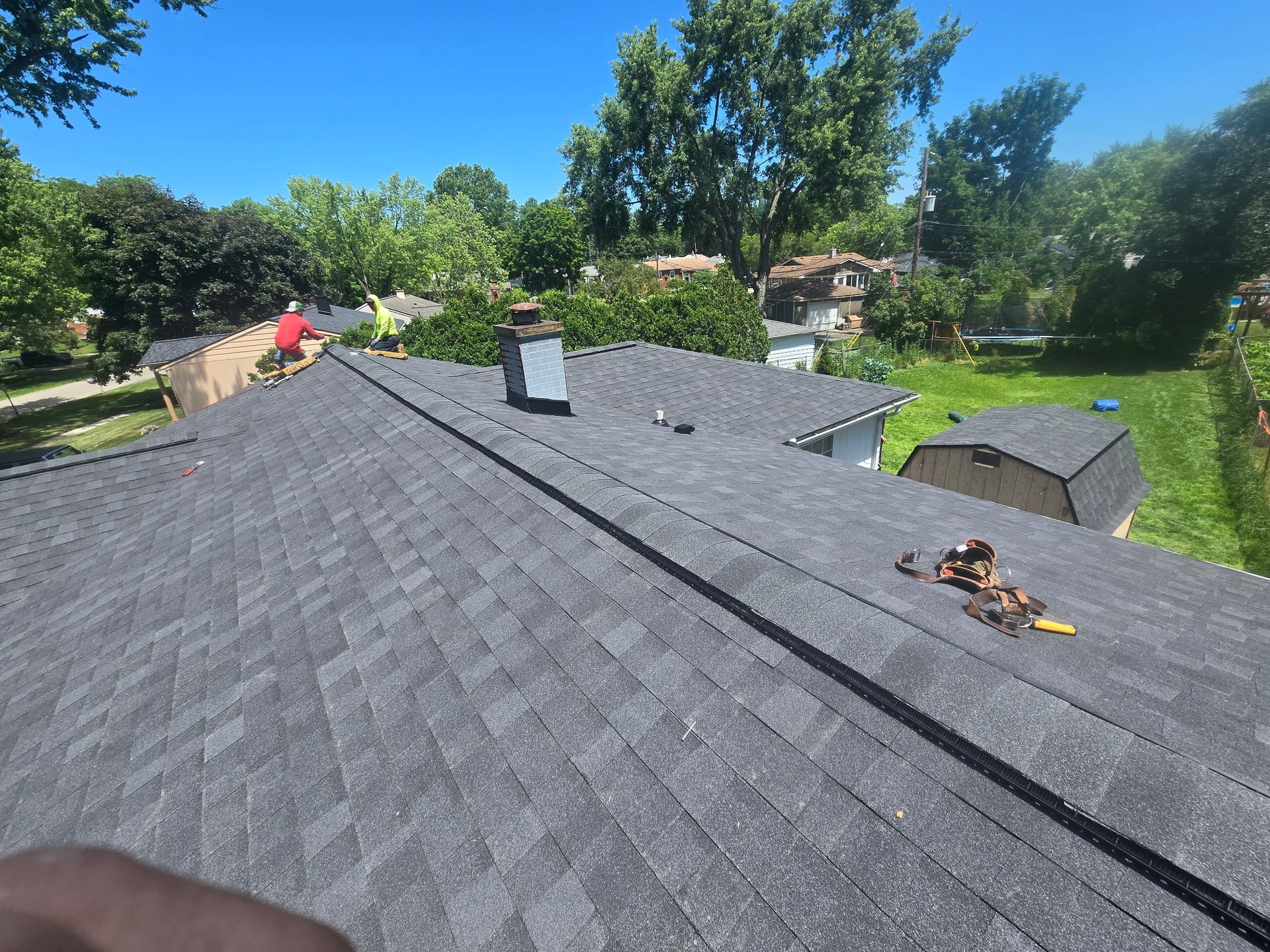 Roofing workers installing dark gray shingles on a house roof in a sunny suburban neighborhood.