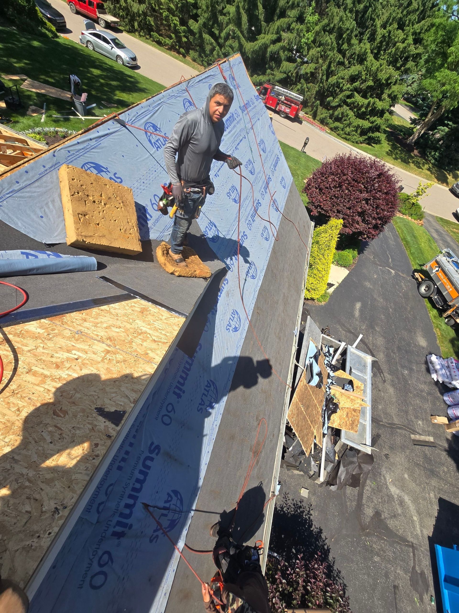 Worker on a steep shingled roof, installing blue underlayment beside rooftop vents and stacked materials. Worker on a steep shingled roof, installing blue underlayment beside rooftop vents and stacked materials.