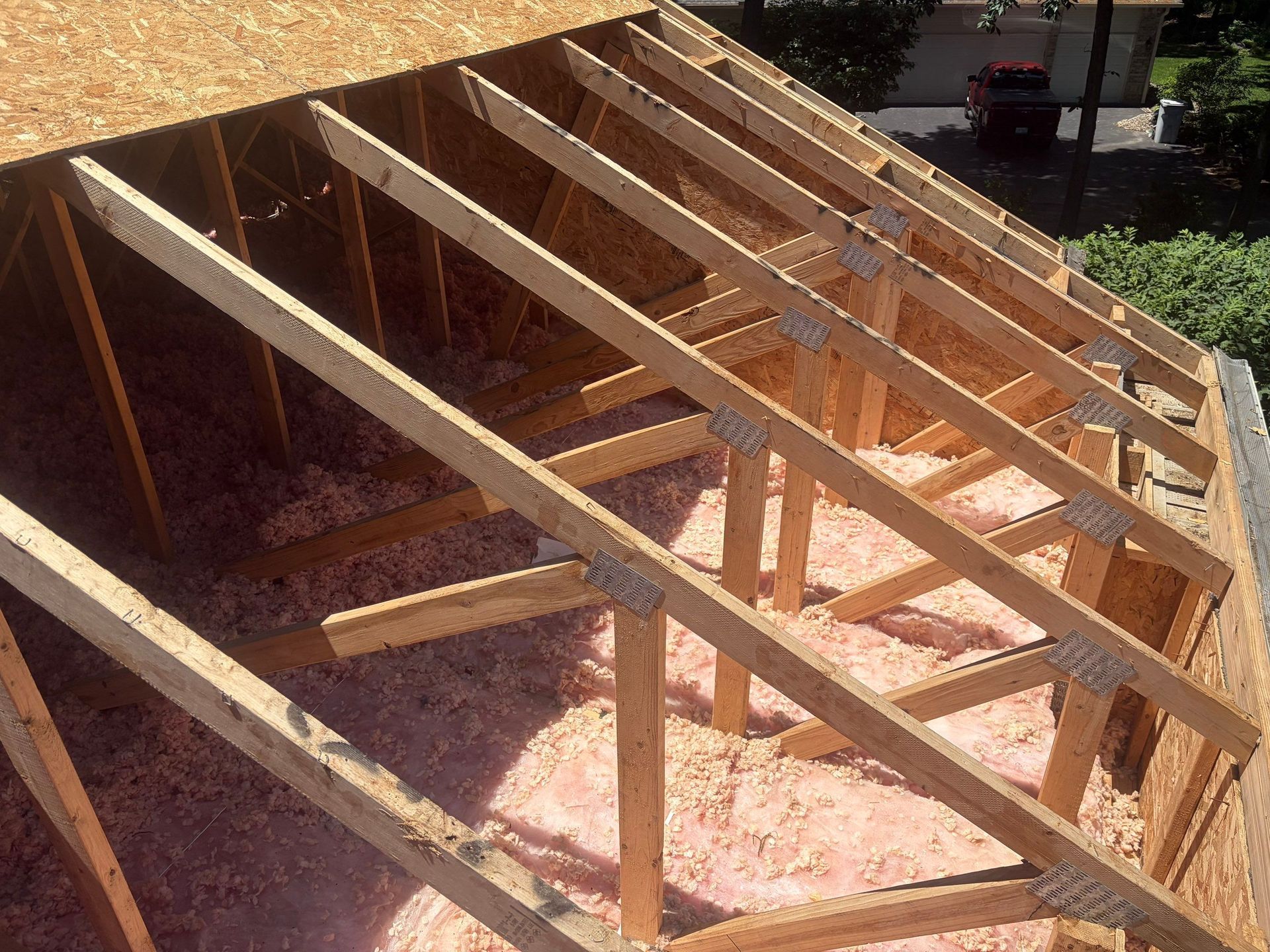 Wooden roof frame under construction beside a house, with beams set over pink insulation.
