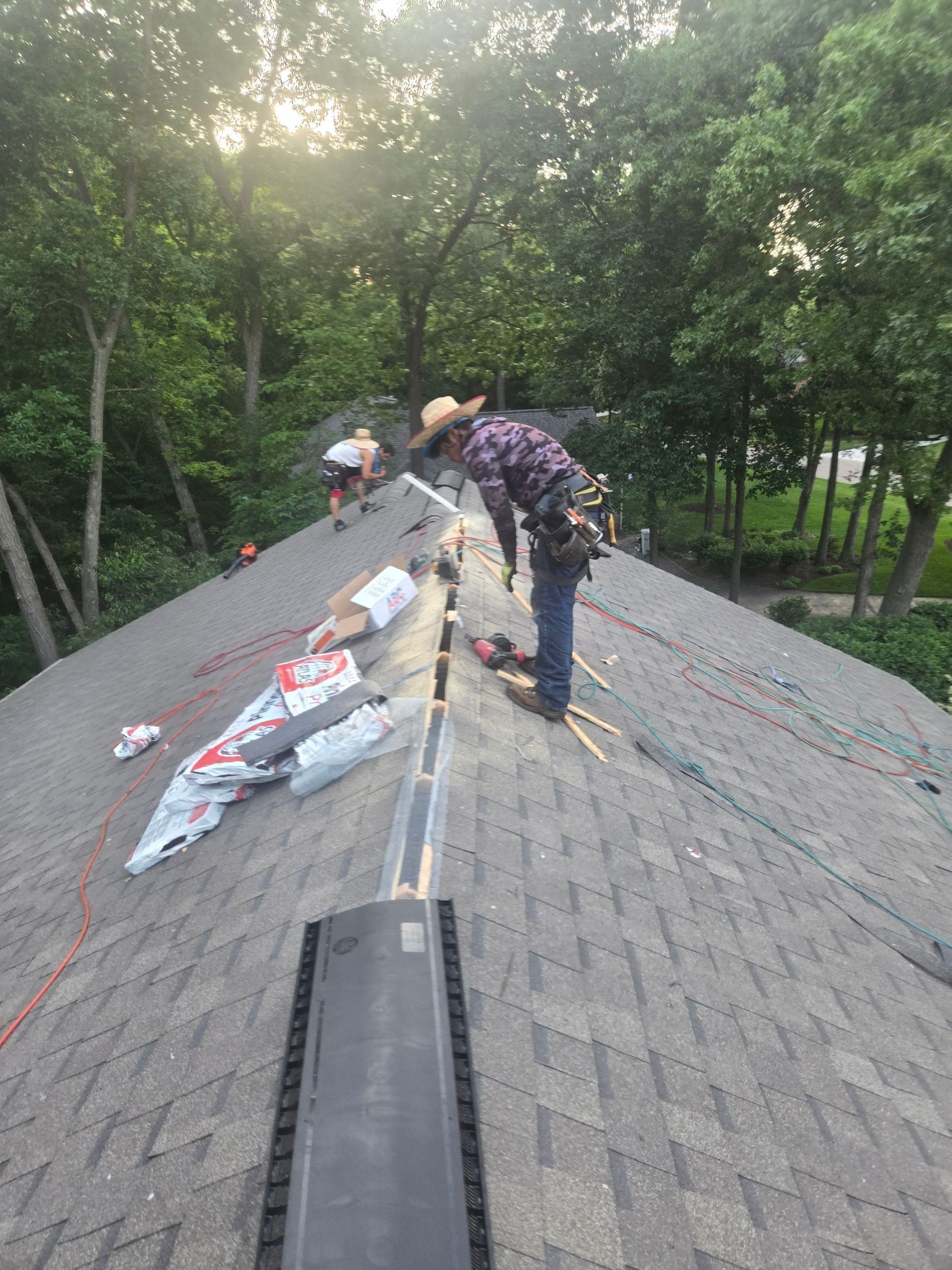Workers repairing shingles on a sloped roof beside trees in daylight Workers repairing shingles on a sloped roof beside trees in daylight
