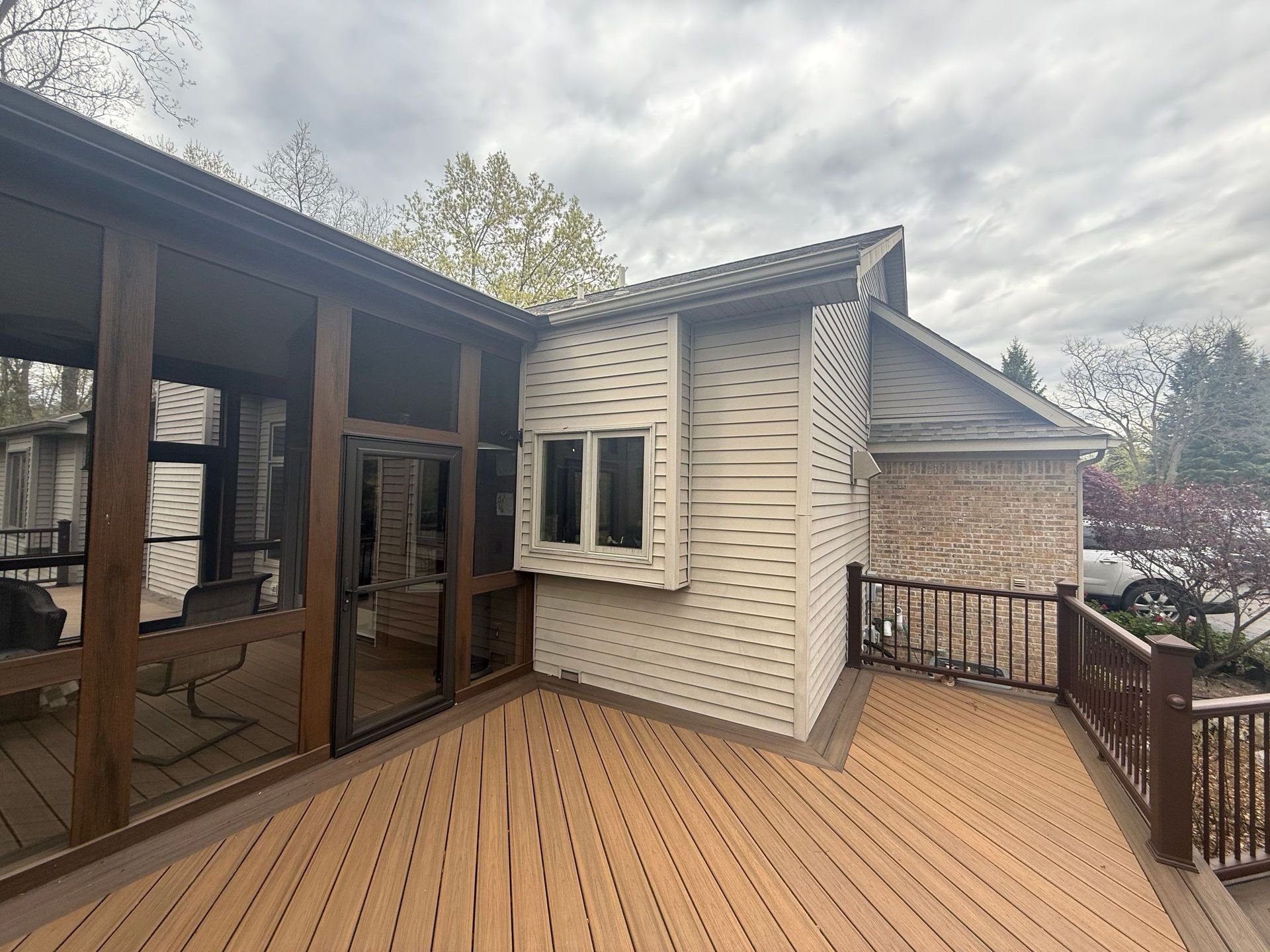Wooden deck beside a house with sliding glass doors, white siding, and a railing overlooking trees and cloudy sky Wooden deck beside a house with sliding glass doors, white siding, and a railing overlooking trees and cloudy sky
