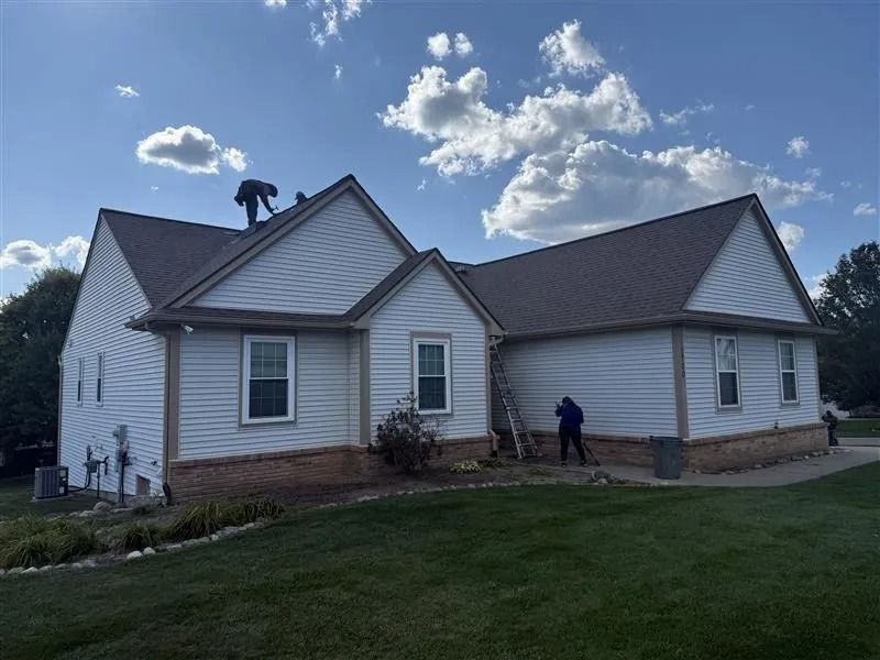 Roofing professionals installing shingles on a light-colored house with a multi-gable roof.