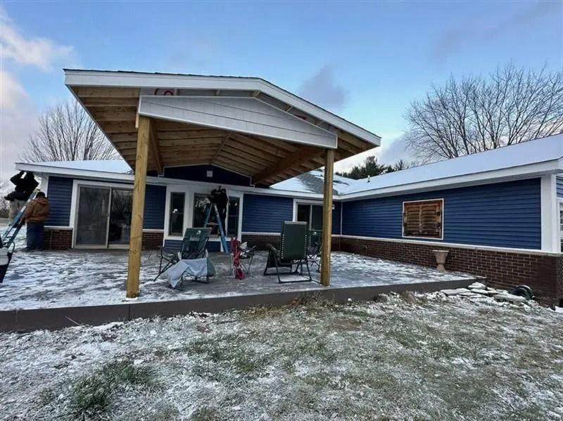 A modern covered patio with exposed wooden beams beside a blue house, with light snow on the ground.