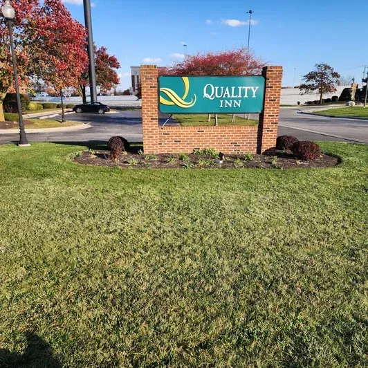Quality Inn roadside sign on a brick base beside a grassy lawn and autumn trees.
