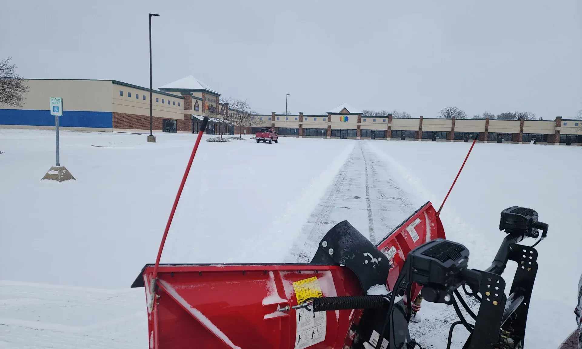 A person wearing a dark coat and gloves clears a snowy sidewalk with a shovel on a sunny winter day.