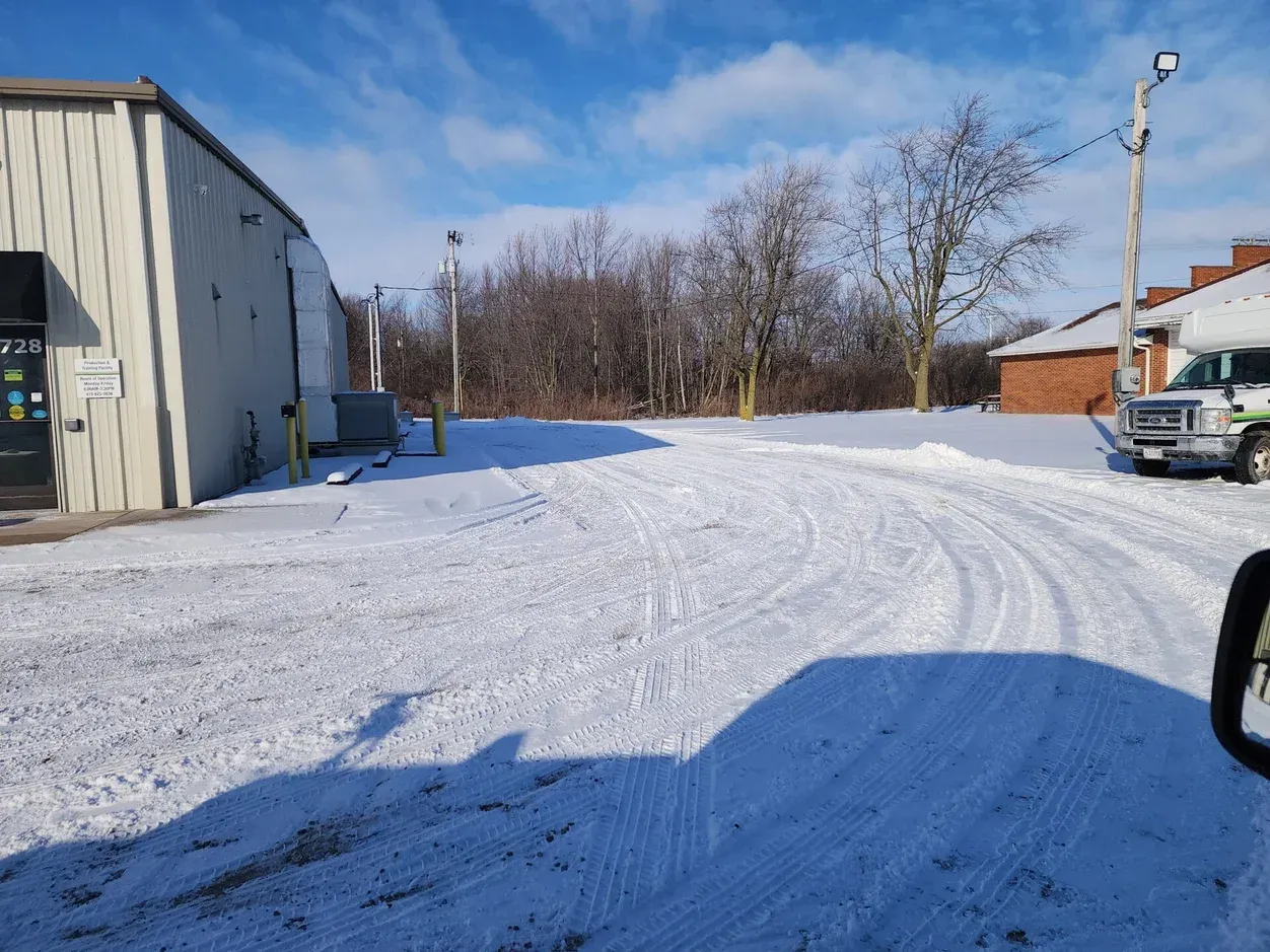 Snowy parking lot beside a gray building on a clear day, with trees and parked vehicles in the distance.