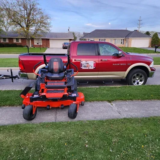 Red pickup truck parked on a suburban street beside an orange zero-turn lawn mower on a trailer.