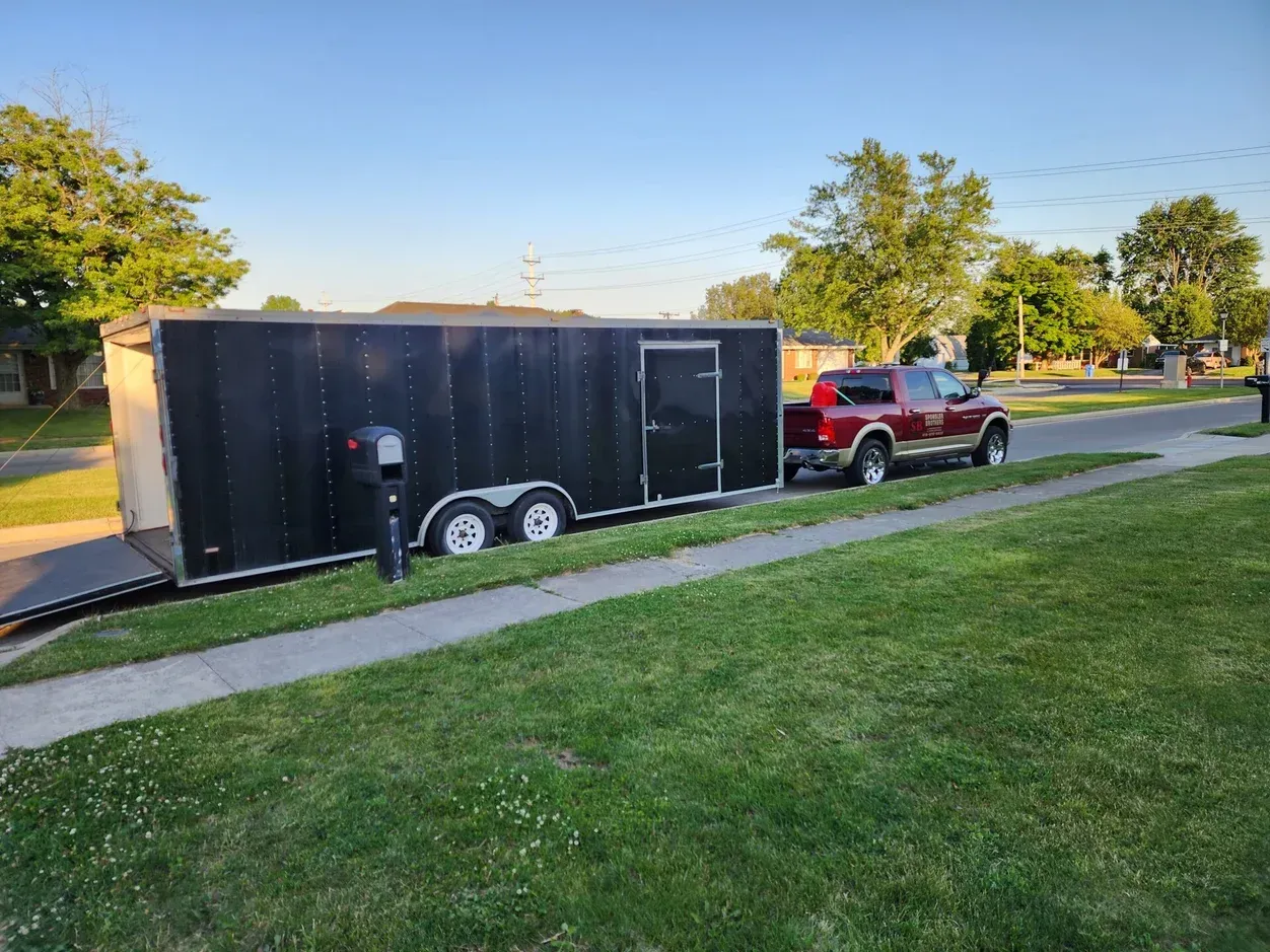 Red pickup towing a large black enclosed trailer along a grassy park path at sunset