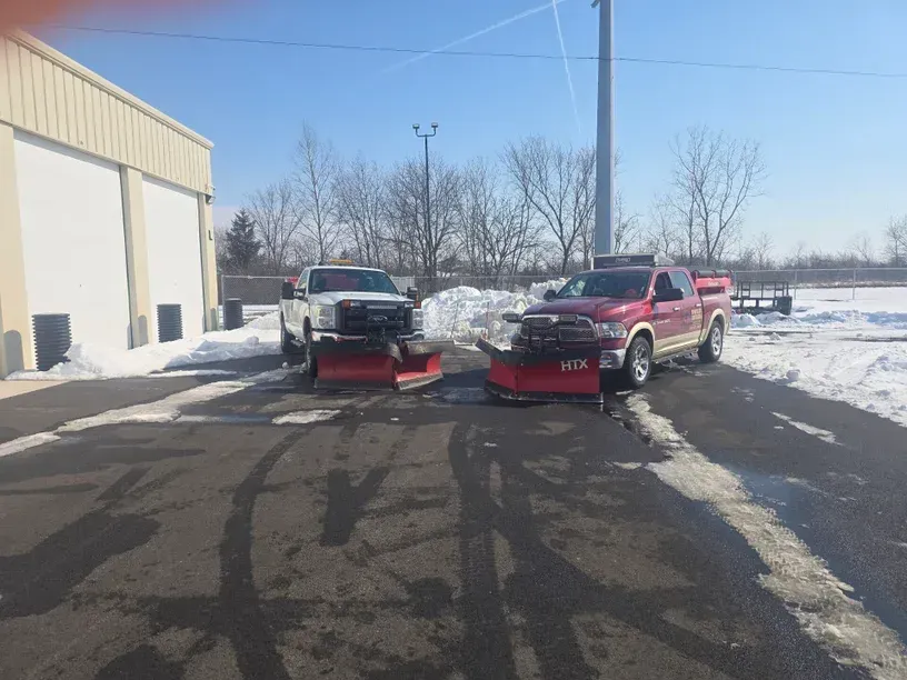 Two pickup trucks with snowplows parked on a snowy lot beside a building