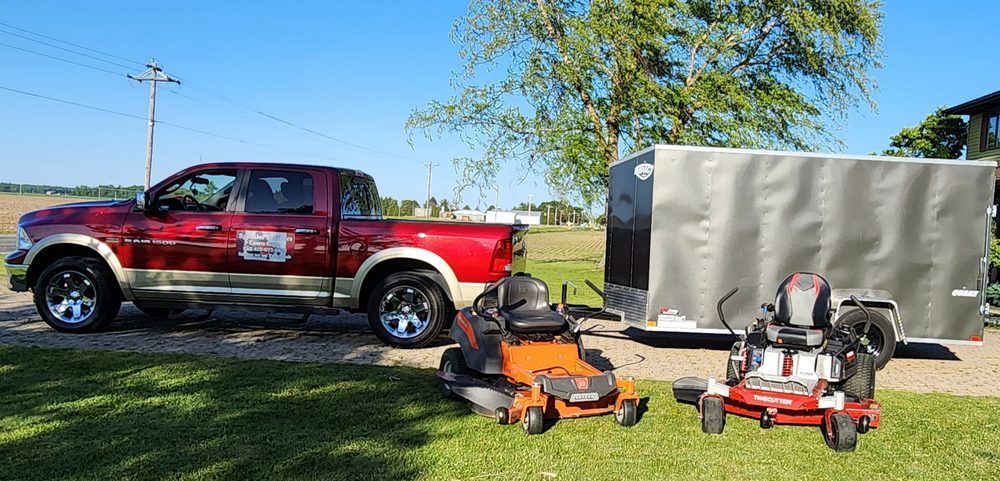 Red pickup truck beside an enclosed trailer and several orange lawn tools on a grassy yard