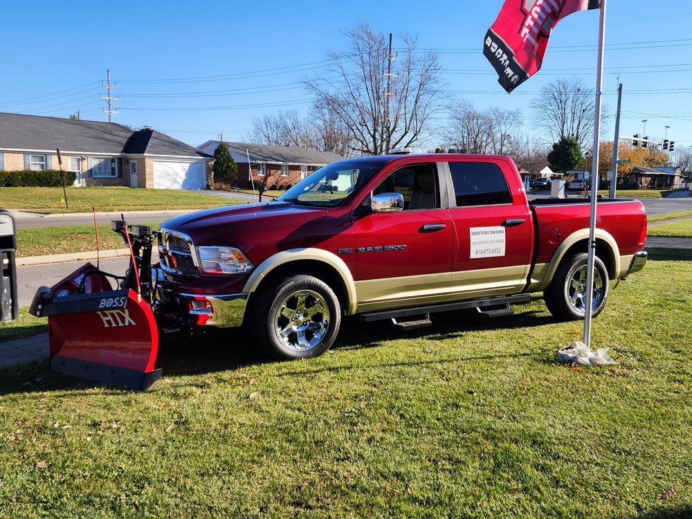Red pickup truck with plow attachment parked on grass beside a flagpole in a sunny residential area