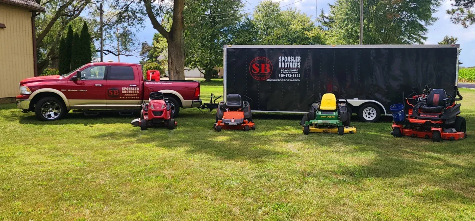 Red pickup truck and four lawn mowers parked on grass beside a black trailer in a sunny yard