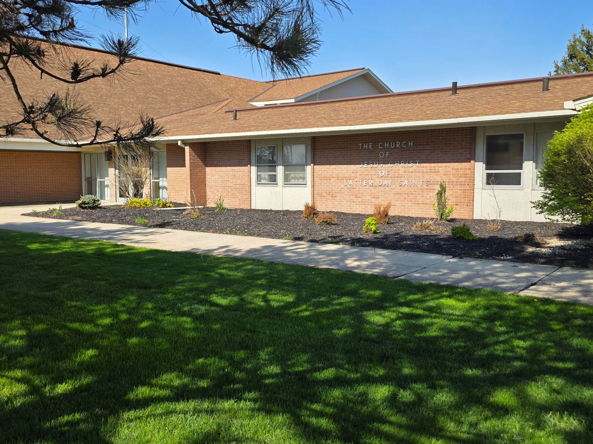 Single-story brick house with a tan roof, front lawn, and shaded walkway under trees