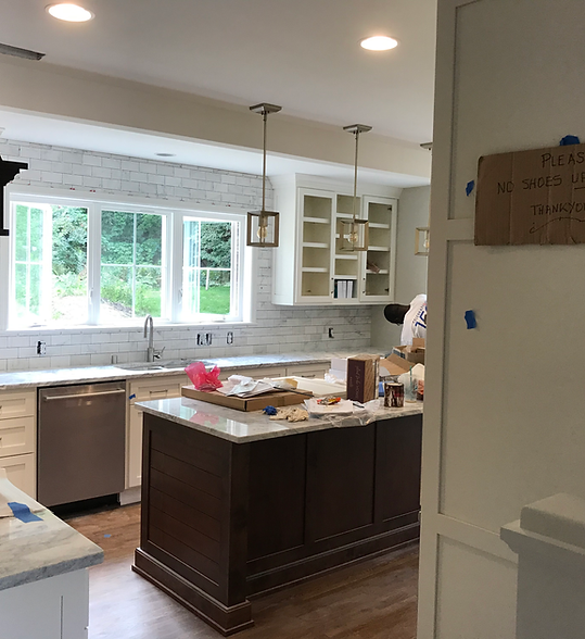 Bright kitchen with dark island, white cabinets, large windows, and an open countertop cluttered with renovation items