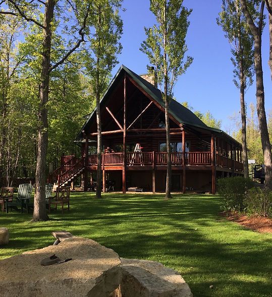 Rustic cabin with a wraparound porch in a sunny wooded yard, framed by tall trees and green lawn