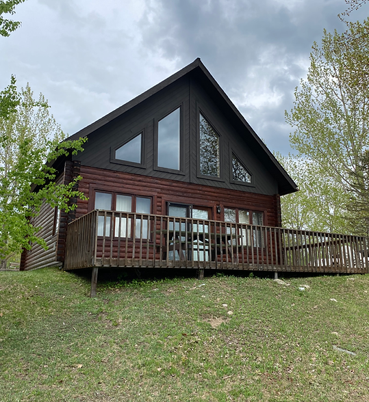 Modern dark-roofed cabin with wood siding and a wraparound deck on a grassy hillside under cloudy sky