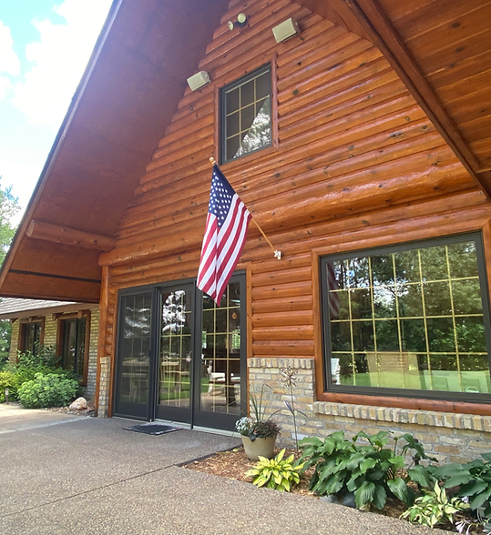 Log cabin-style building entrance with American flag, large windows, and stone planter border