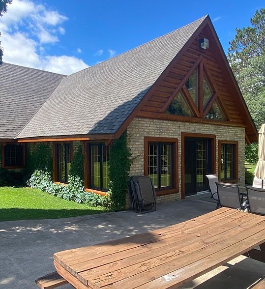 Wooden cabin-style house with tall peaked roof, large windows, and a sunlit patio with outdoor furniture