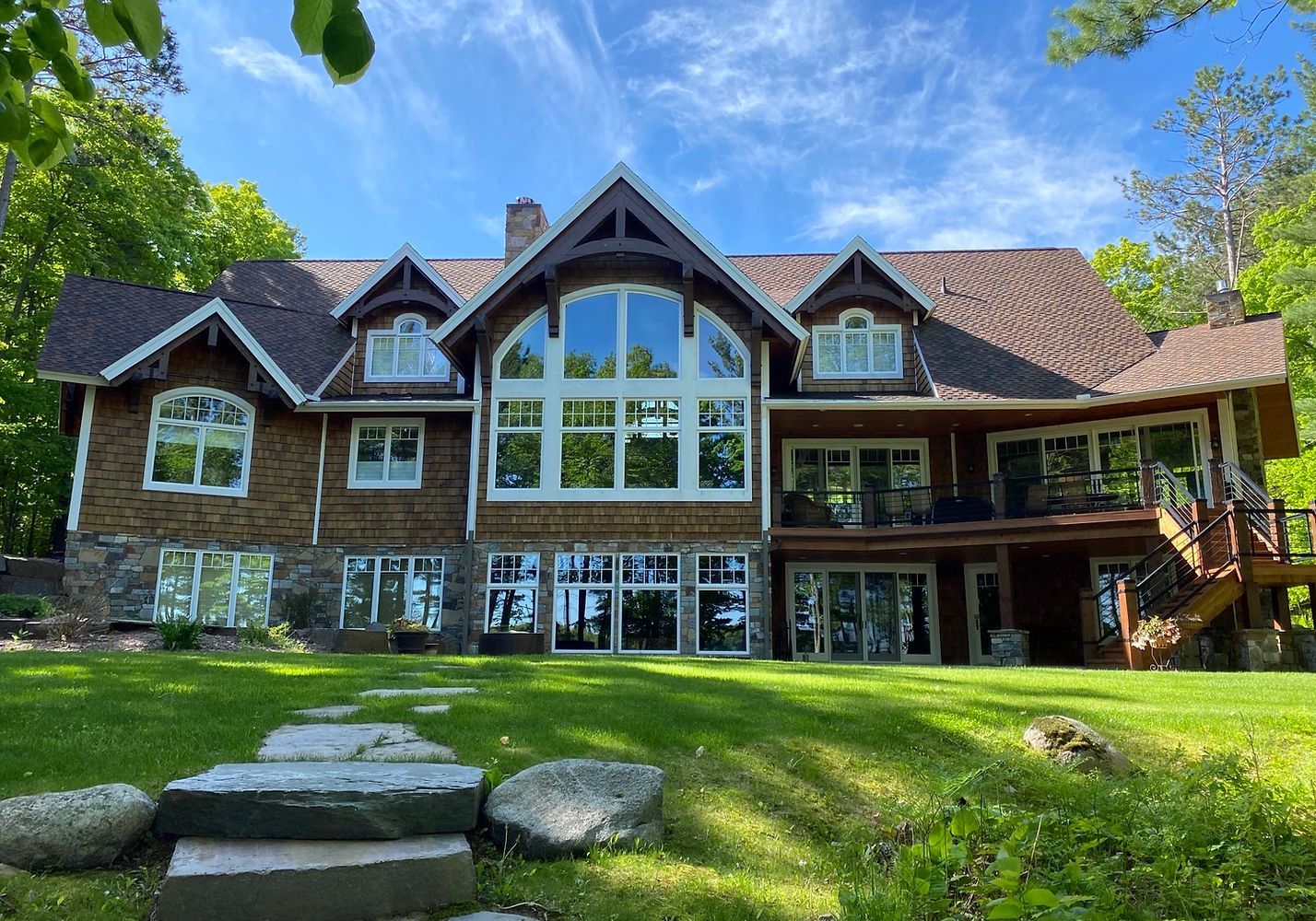 Large rustic house with stone and wood exterior, big central windows, and a green lawn under blue sky