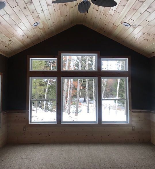 Empty cabin interior with wood ceiling, dark walls, and large windows overlooking snowy trees