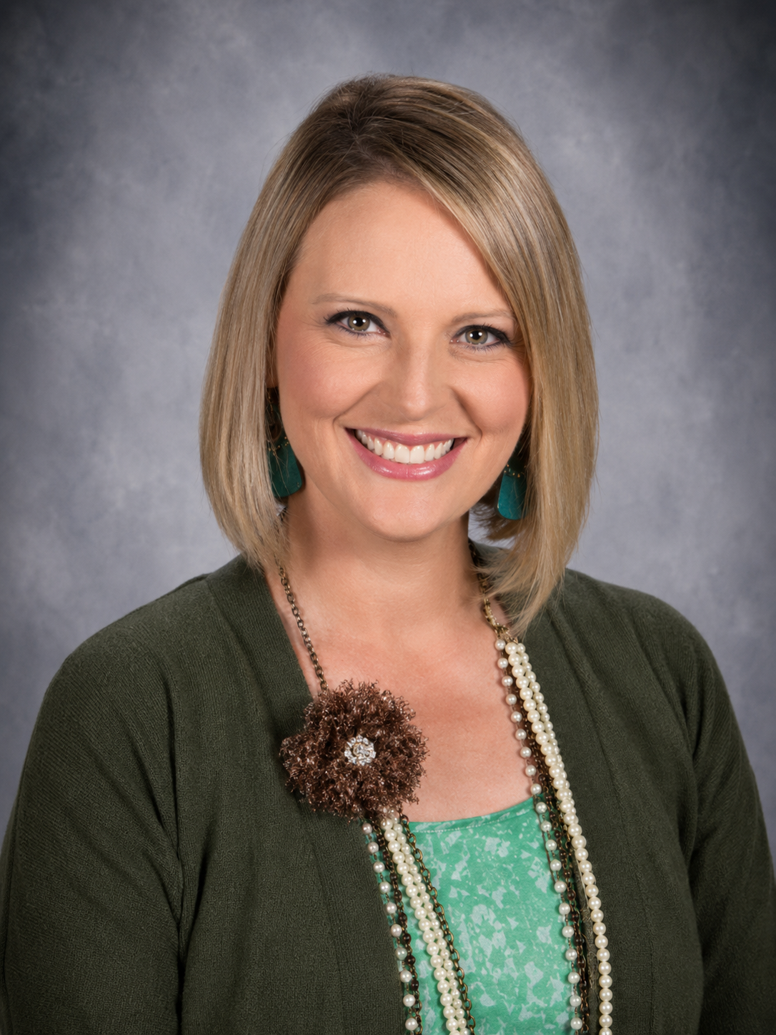 Blonde woman smiles, wearing a necklace and a blue cardigan over a light blue shirt.