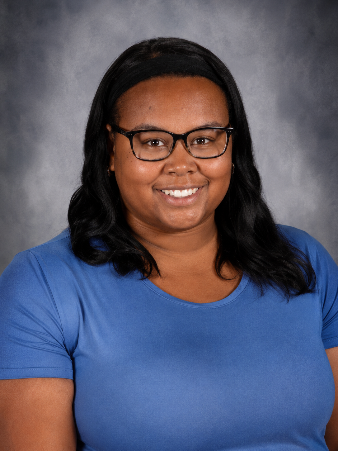 Woman with glasses and braids smiles, wearing a floral blouse, against a blurred backdrop.