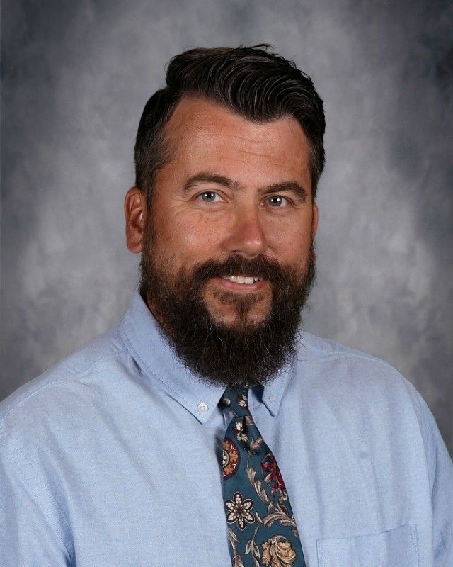 Man with beard, wearing a sweater and button-down shirt, smiling at the camera against a blurred background.