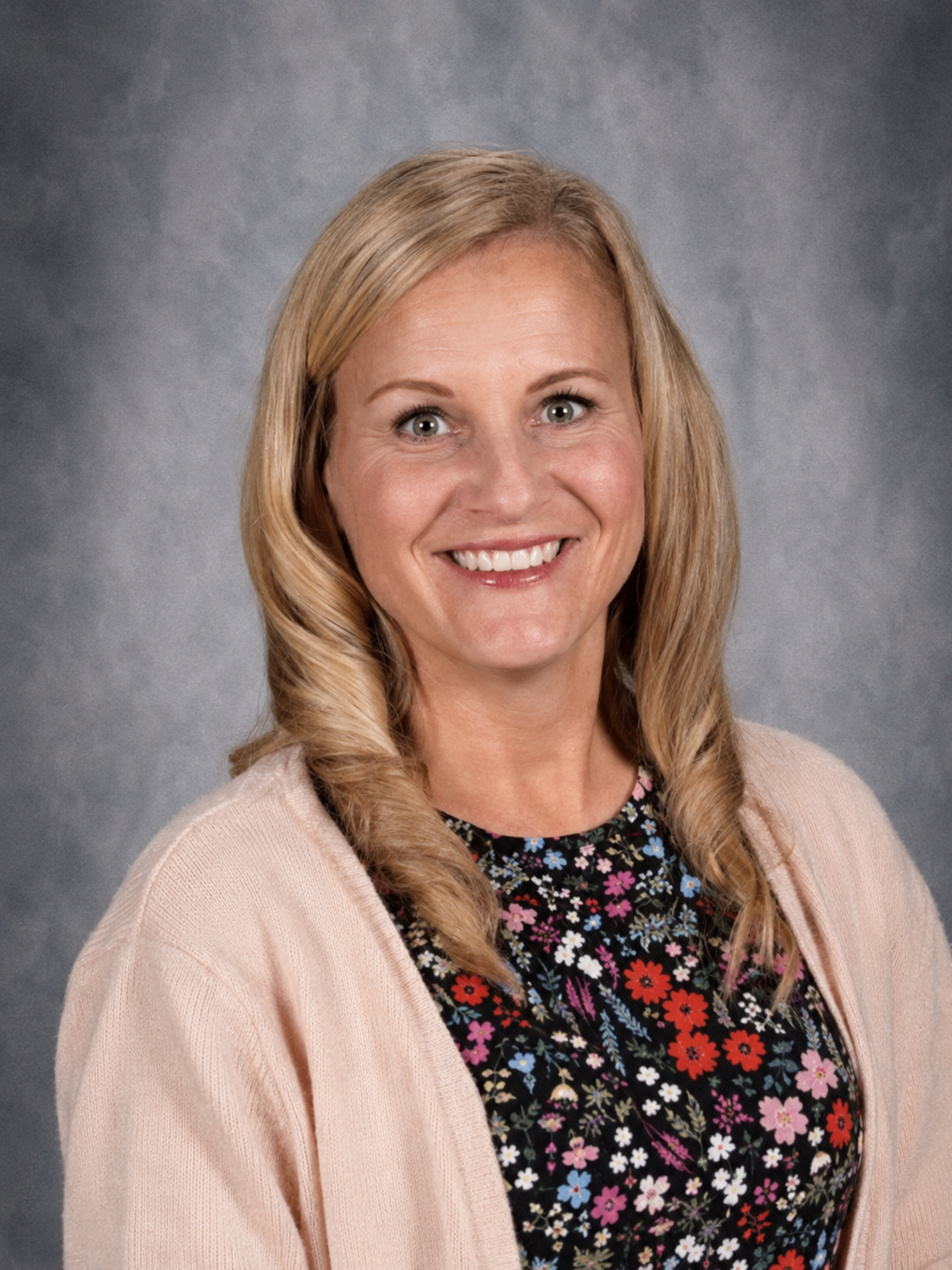Blond woman with a bright smile, wearing a gray sweater and colorful beaded necklace, posing for a photo.