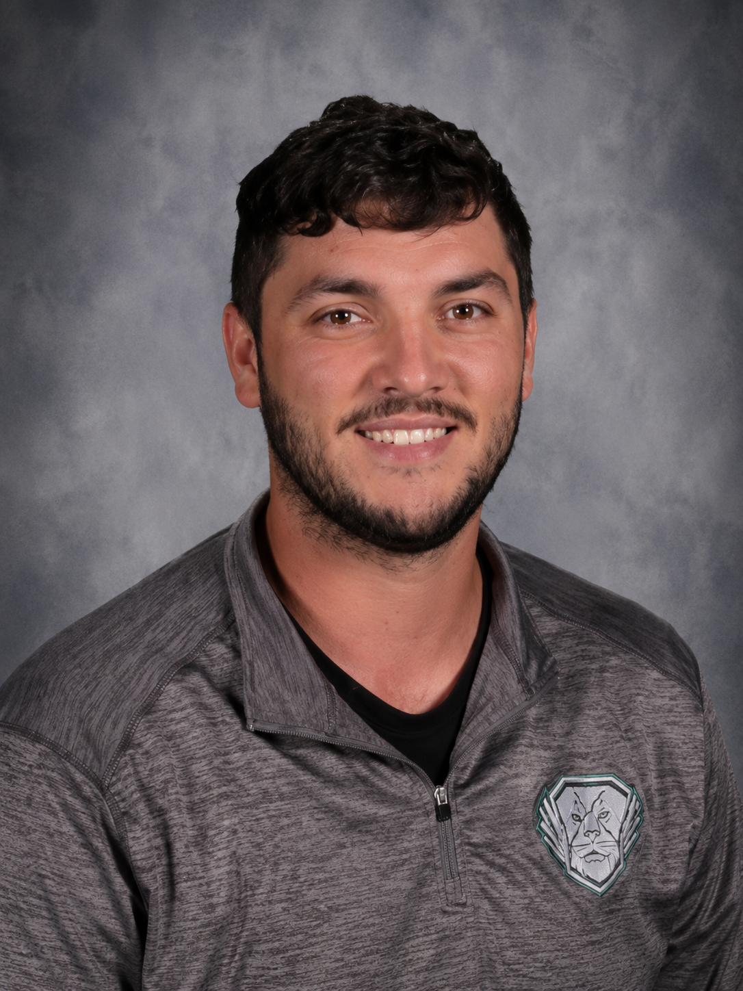 Smiling man with a full beard, wearing a brown polo shirt against a mottled gray backdrop.