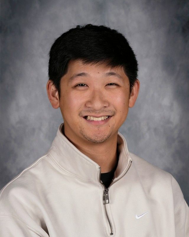 Smiling Asian man in a black hooded jacket, against a blurred gray background.