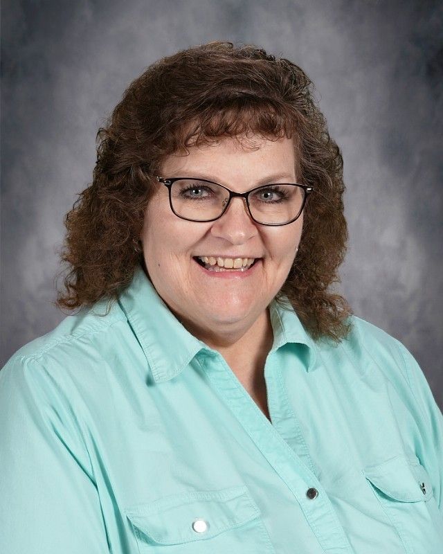 Woman with glasses, brown curly hair, and gray shirt, against a blurred gray backdrop.