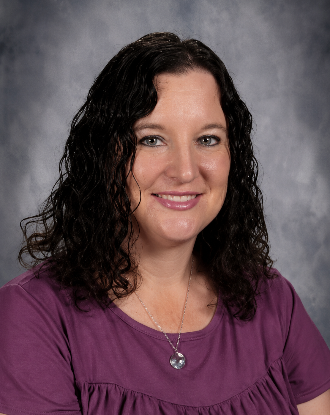 Woman with dark wavy hair, smiling, wearing a red shirt, in front of a grey backdrop.
