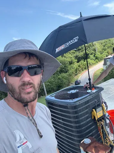 Man in a hat and sunglasses on a rooftop with an air conditioning unit, shaded by an umbrella.