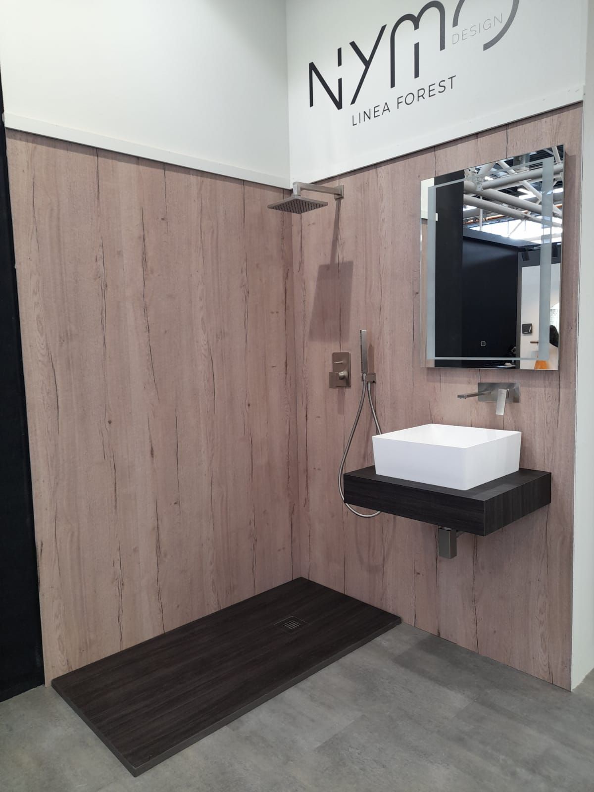 Esposizione del bagno con pannelli in legno, doccia a pioggia, mobile lavabo sospeso con lavandino e specchio.