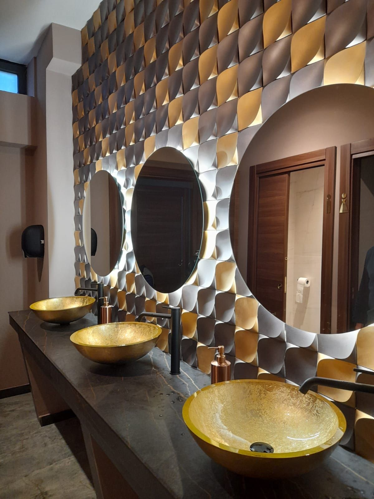 Bagno con tre specchi rotondi, lavandini dorati, parete decorata, bancone scuro e porte.