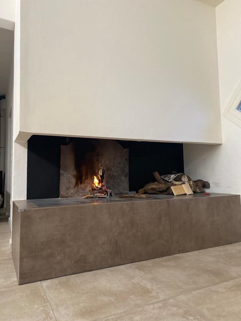 Camino con fiamme e ceppi ardenti; interno scuro, focolare in pietra, parete bianca.