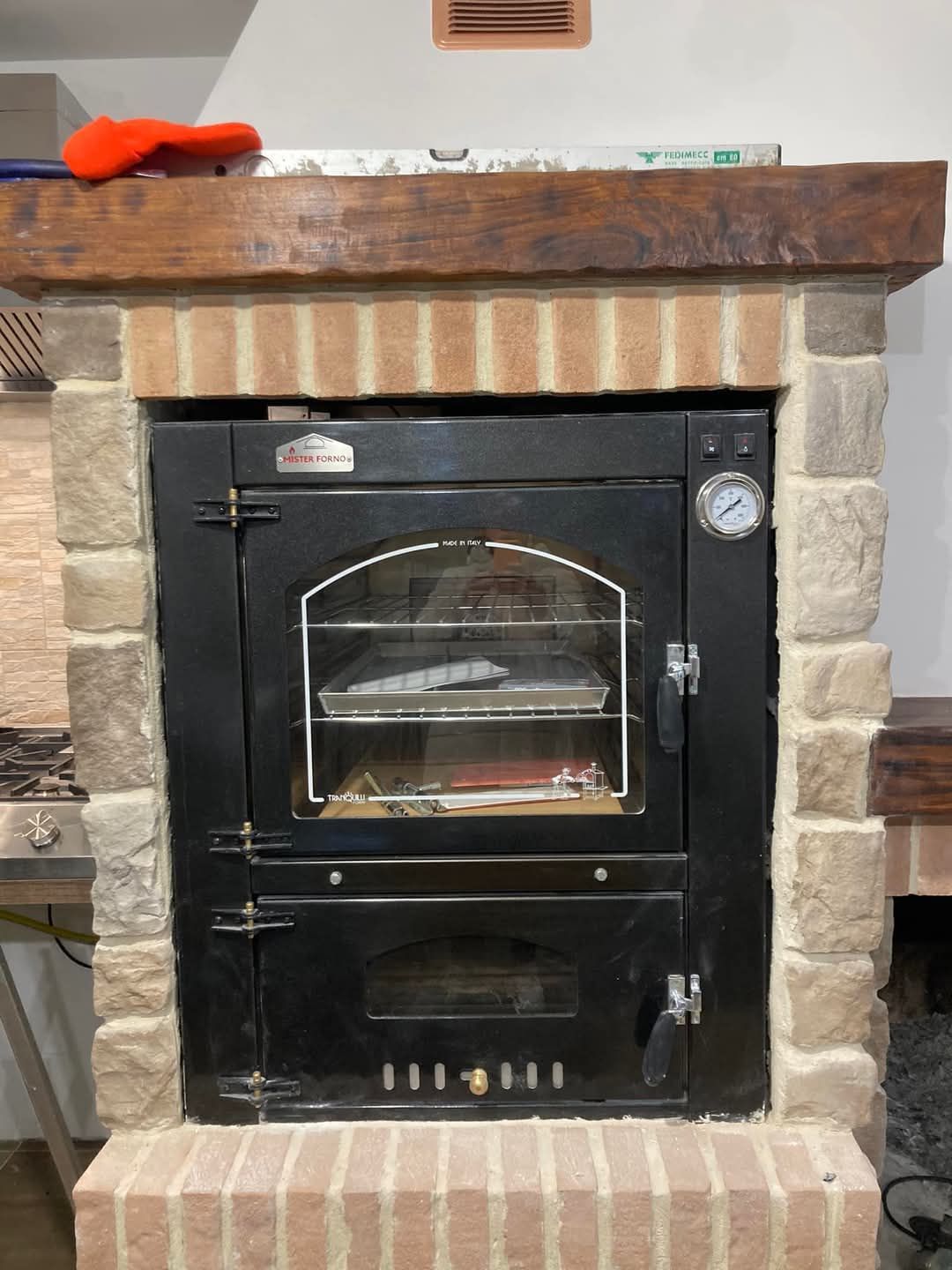Forno nero incassato in una struttura in mattoni e pietra, con sportello in vetro e indicatore della temperatura.