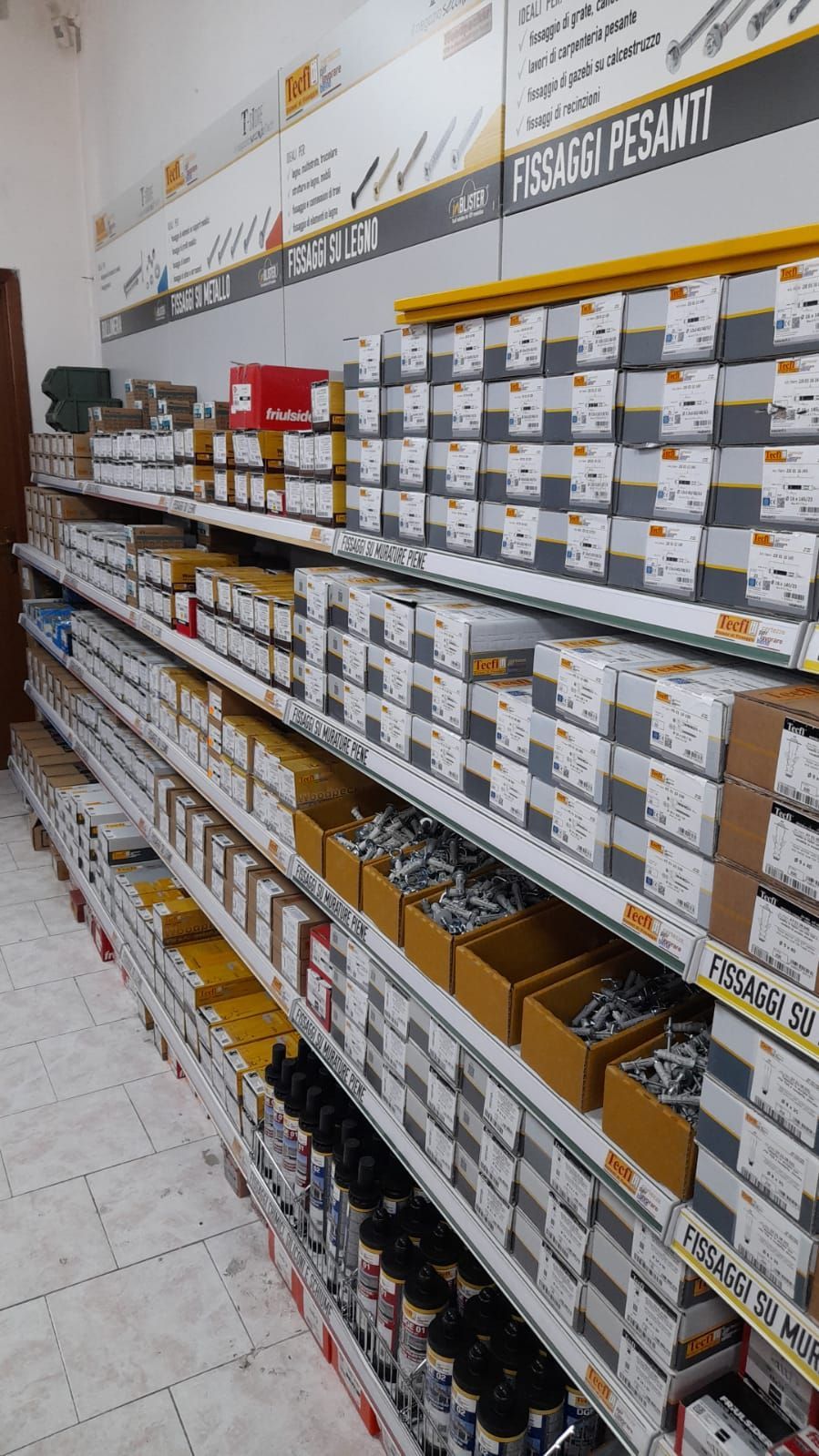 Scaffali pieni di materiale di ferramenta in un negozio; scatole, contenitori e prodotti etichettati.